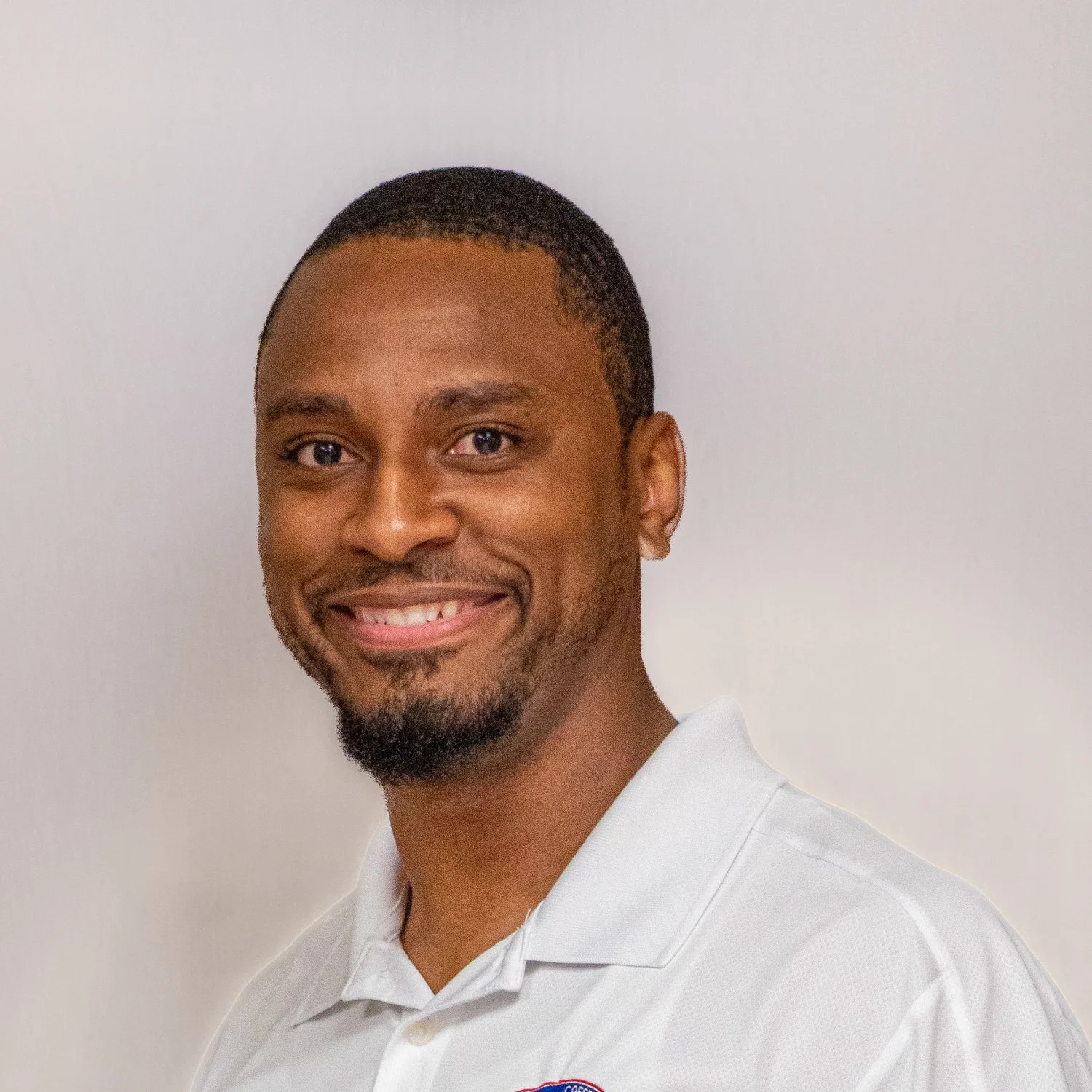 Portrait of a smiling man with short hair and a beard, wearing a white collared shirt with a small logo on the chest, against a plain light-colored background.