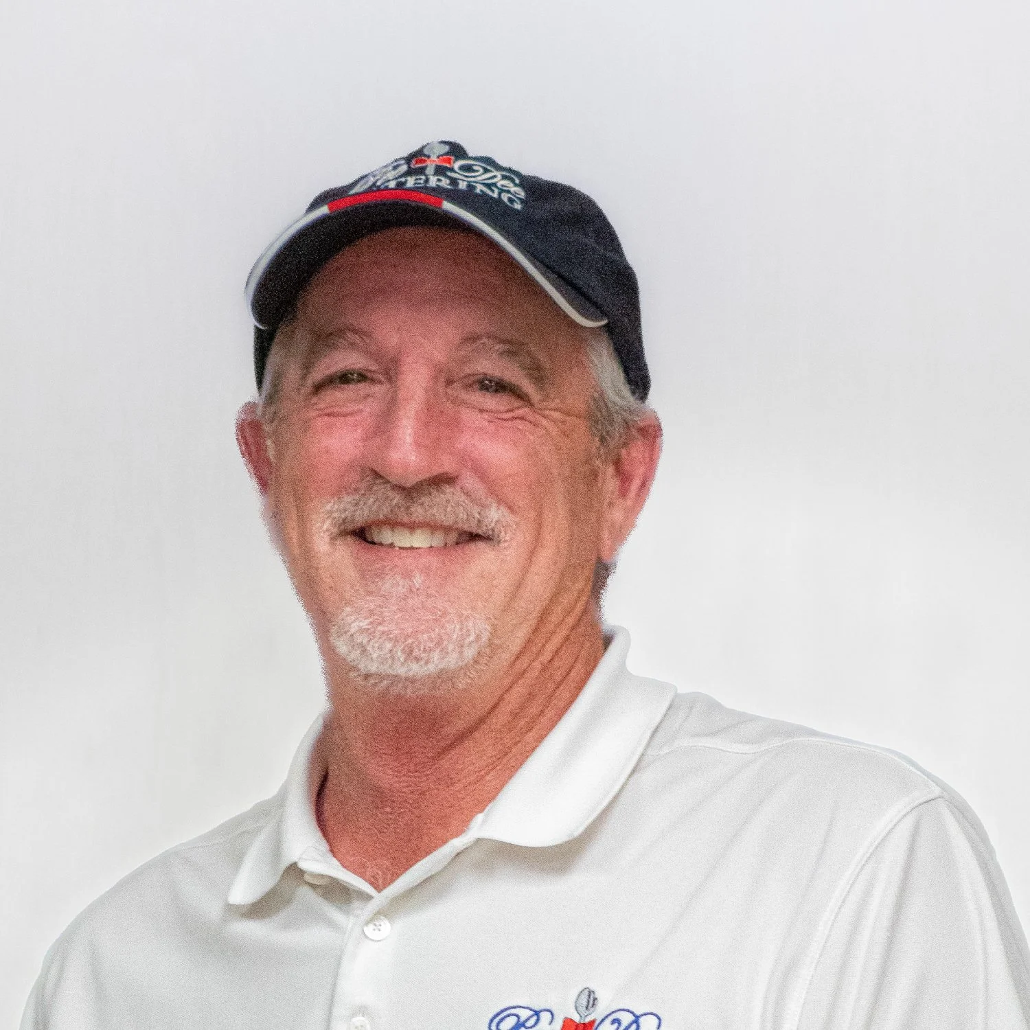 A middle-aged man with a white beard and mustache smiling, wearing a black cap with a logo and white polo shirt with a small logo.
