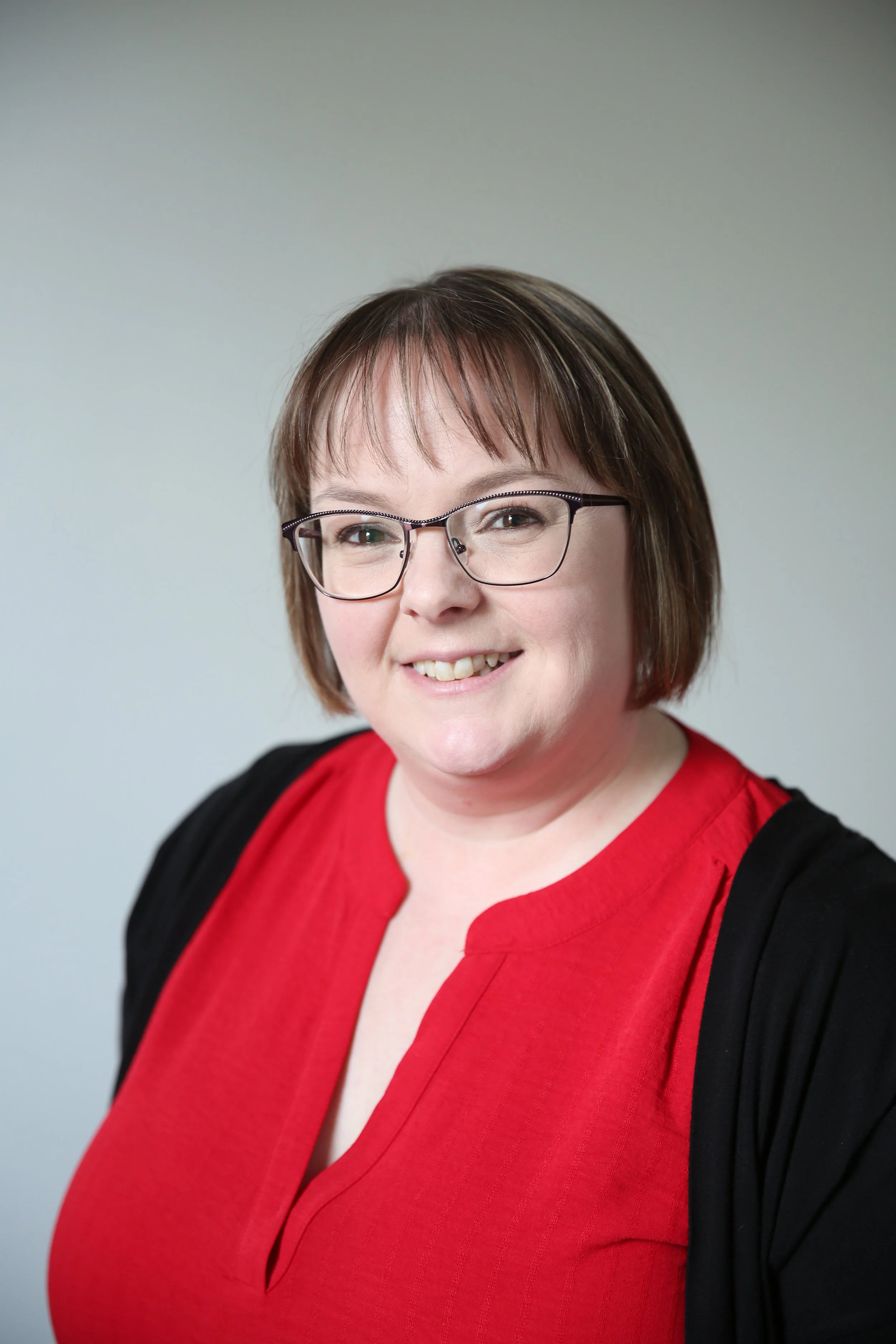 Portrait of a woman with short brown hair, glasses, wearing a red top and a black cardigan, smiling against a plain light gray background.