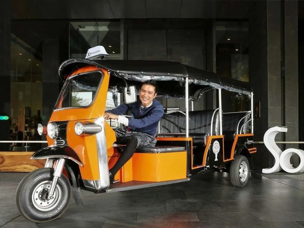 A smiling man in a suit driving a bright orange tuk-tuk in an indoor setting, with a large white letter 'S' next to it.
