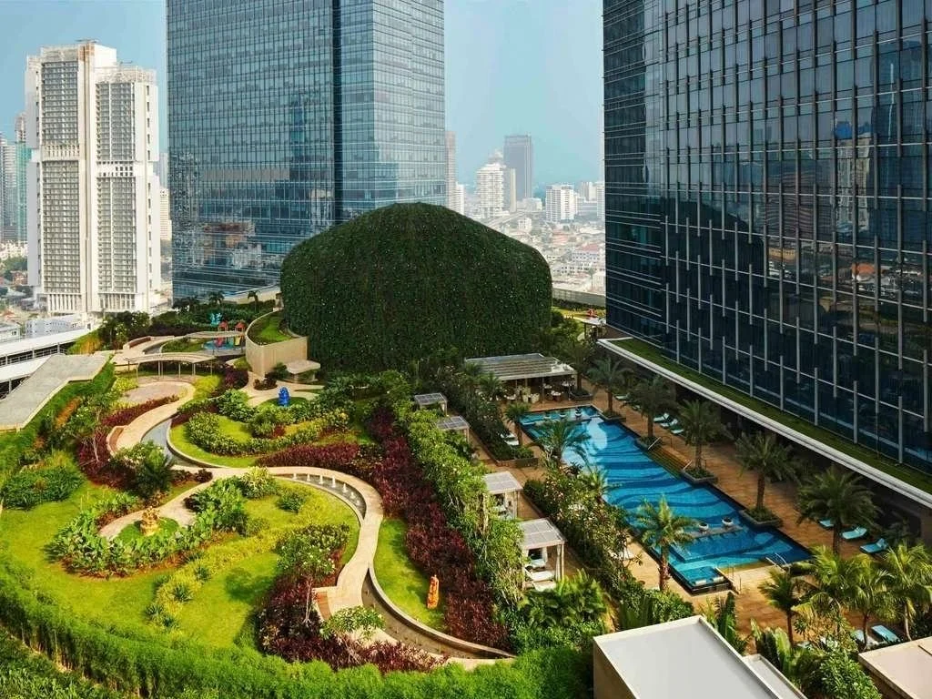 A rooftop garden and pool area surrounded by tall modern office buildings, with green lawns, trees, winding pathways, and a rectangular swimming pool with lounge chairs along the sides.