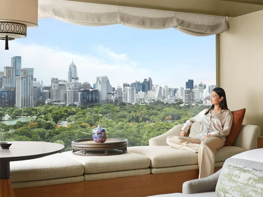 Guest relaxing on an expansive window daybed overlooking Lumphini Park and the Bangkok city skyline in a luxury suite at Dusit Thani Bangkok, Thailand.