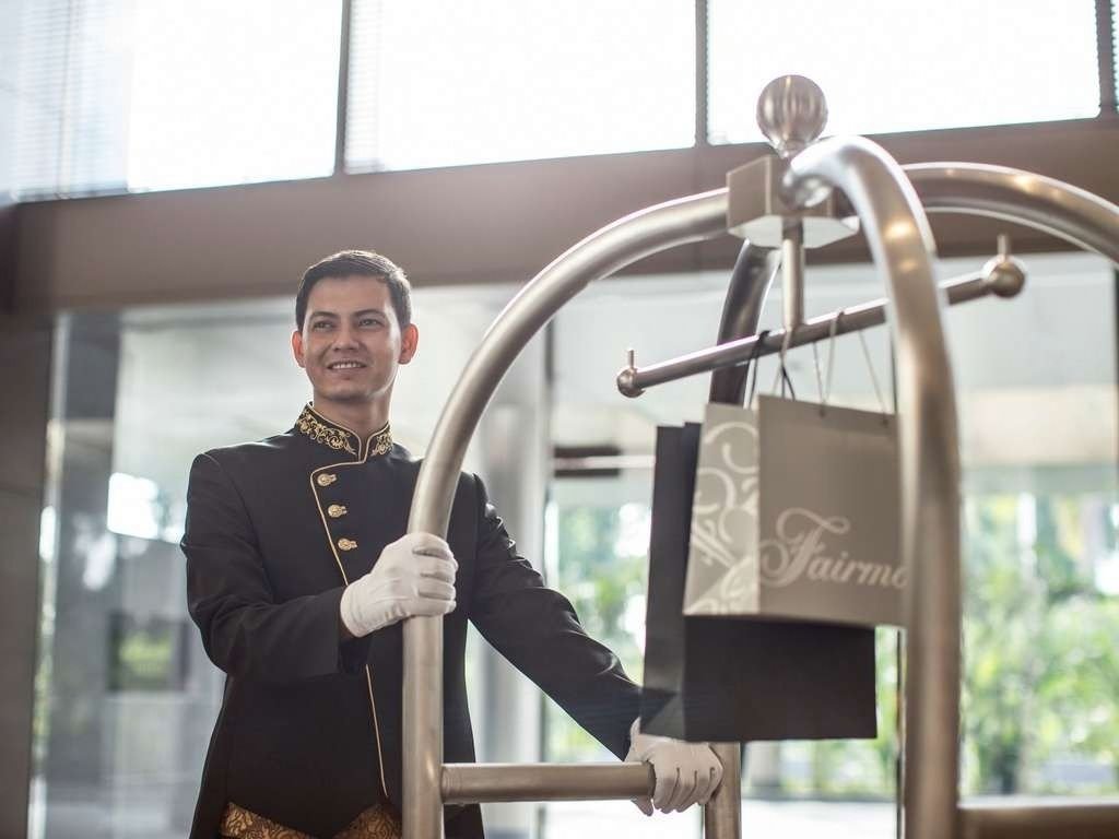 A bellhop in a black uniform with gold trim and white gloves stands at a hotel entrance, holding a luggage cart with shopping bags on it.