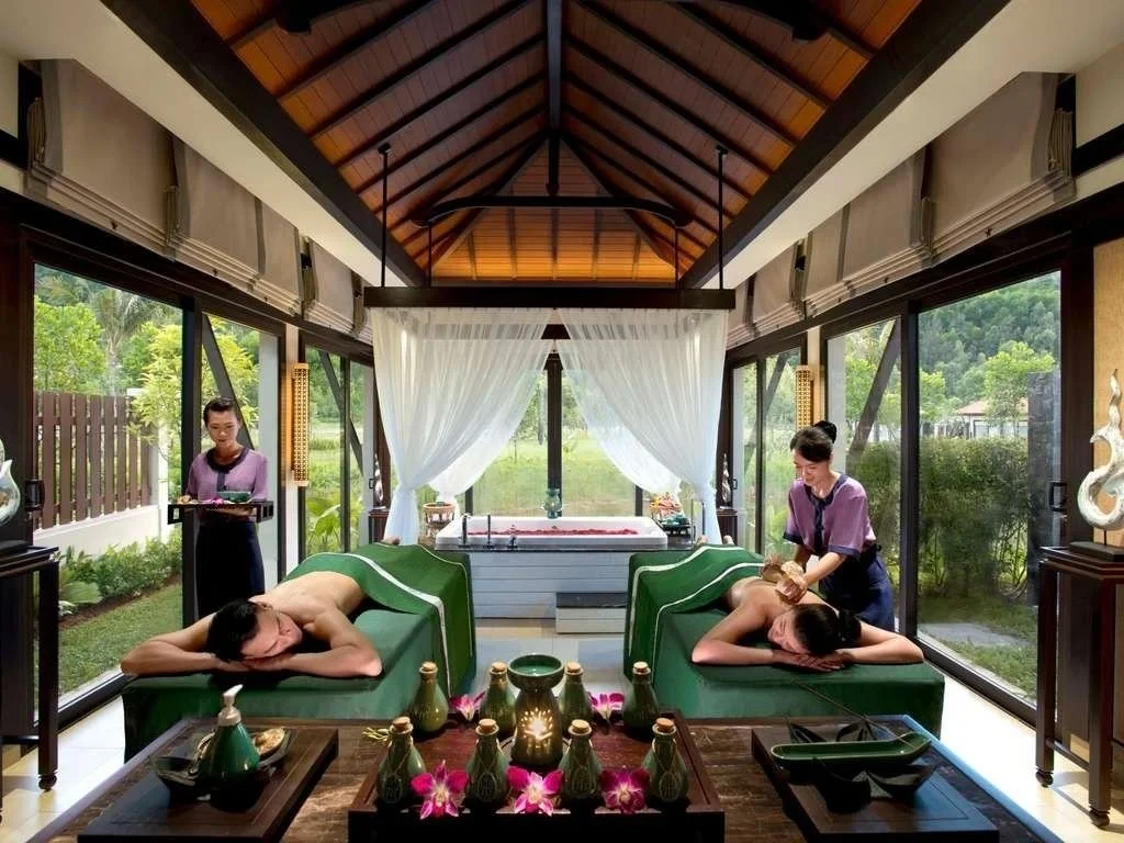 Two women receiving massage treatments at a spa with large windows, outdoor greenery, and a relaxing interior with massage tables, decorative items, and a mountainous landscape outside.