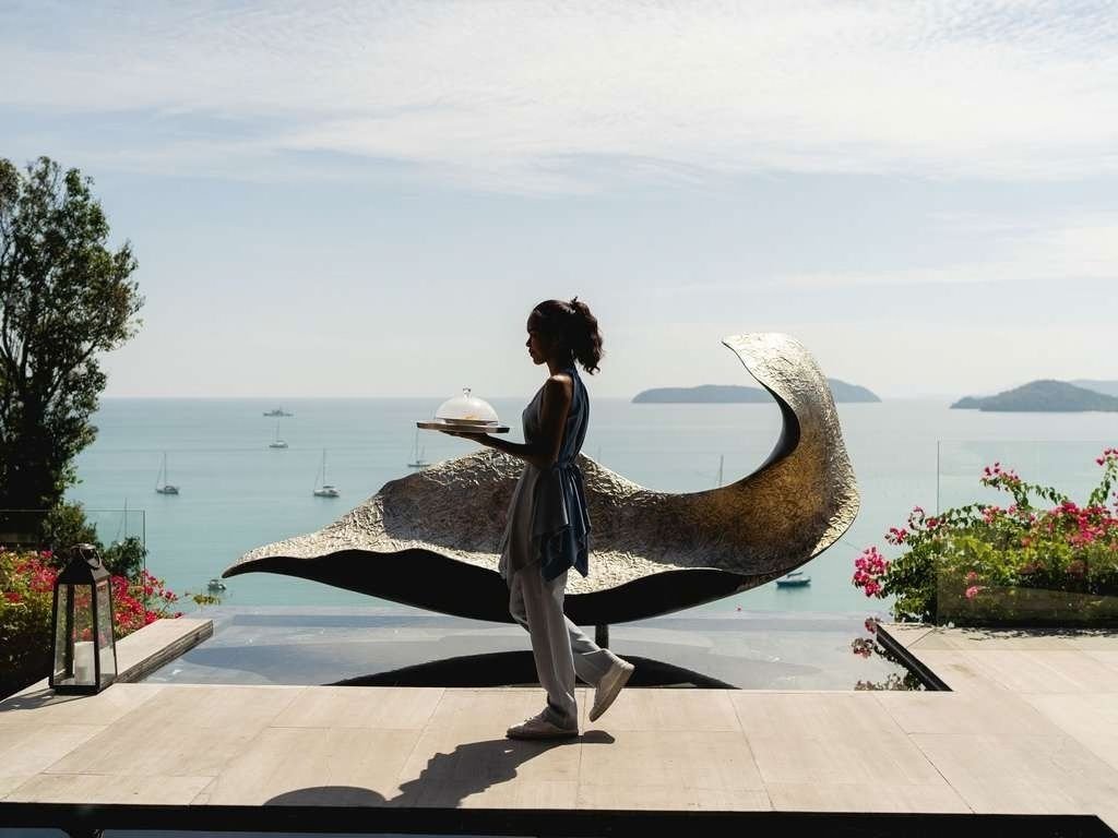 A woman in a dark top and light pants is holding a tray with a covered dish, standing on a terrace overlooking a body of water with boats. There is a large, abstract sculpture behind her, and flowers in planters on either side.
