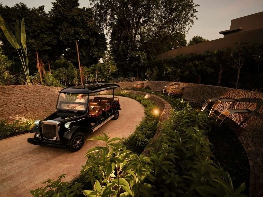 A vintage black and brown golf cart is driving along a winding brick path surrounded by lush greenery and trees, with soft lights illuminating the scene during dusk.