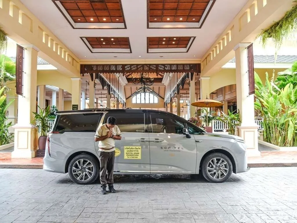 A person is standing near a silver van with the logo Sofitel, holding a phone and looking down, in front of a hotel entrance with lush greenery and wooden decor.