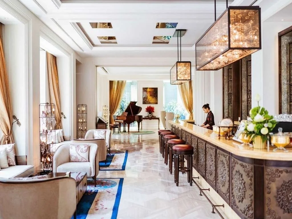 Luxurious hotel lobby with seating area, bar counter, piano, a woman sitting at the bar, chandelier, and large windows with curtains.