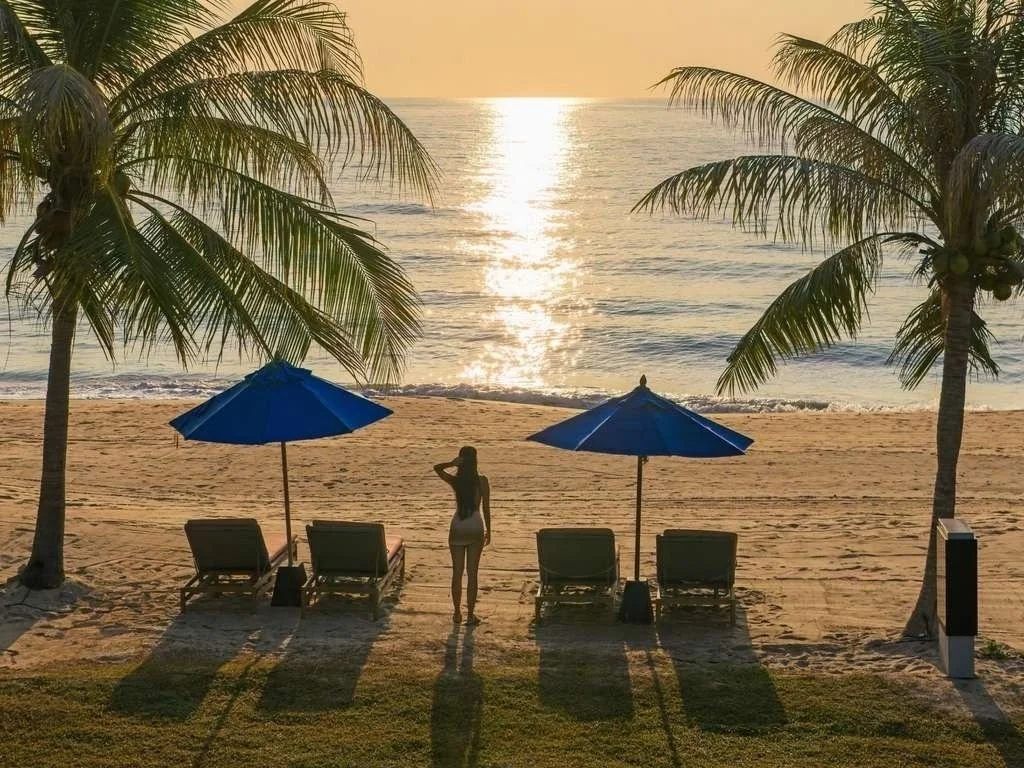 A woman stands on the sandy beach with her back to the camera, facing the ocean at sunset. There are two blue umbrellas and four lounge chairs on the beach, with palm trees on either side.