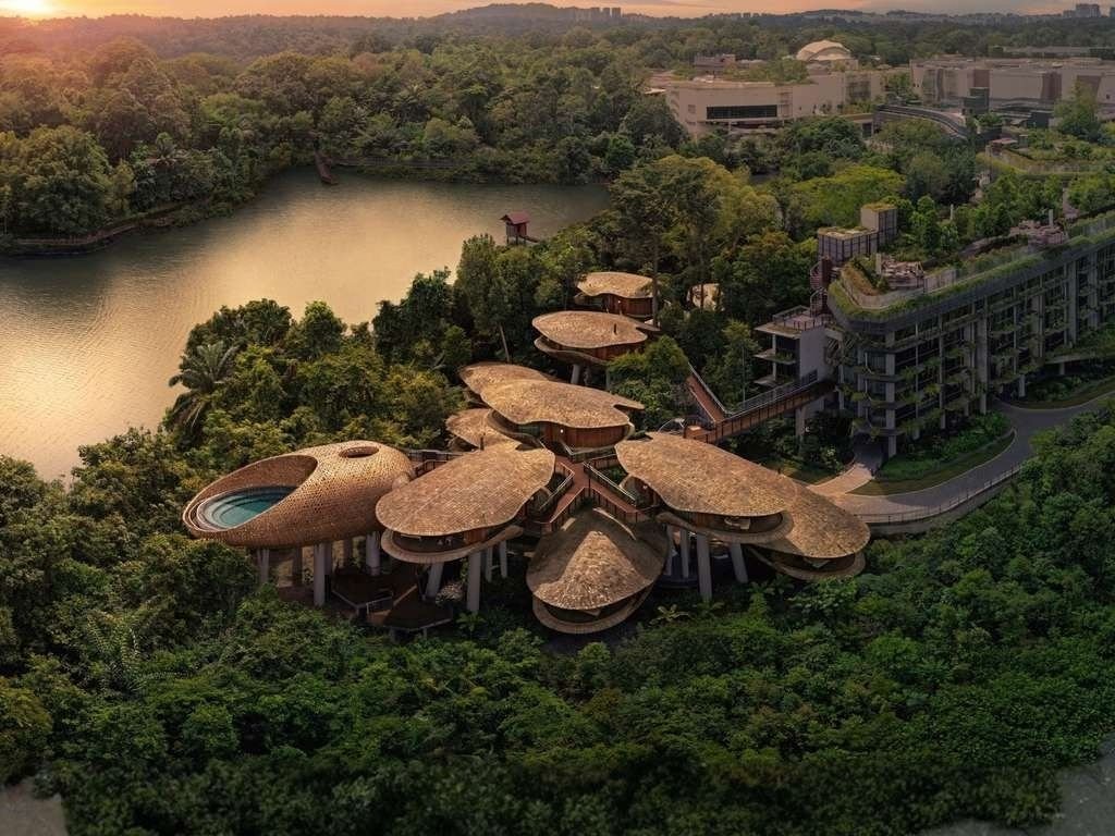 Aerial view of a futuristic building with mushroom-shaped structures surrounded by lush greenery and a lake in the background during sunset.