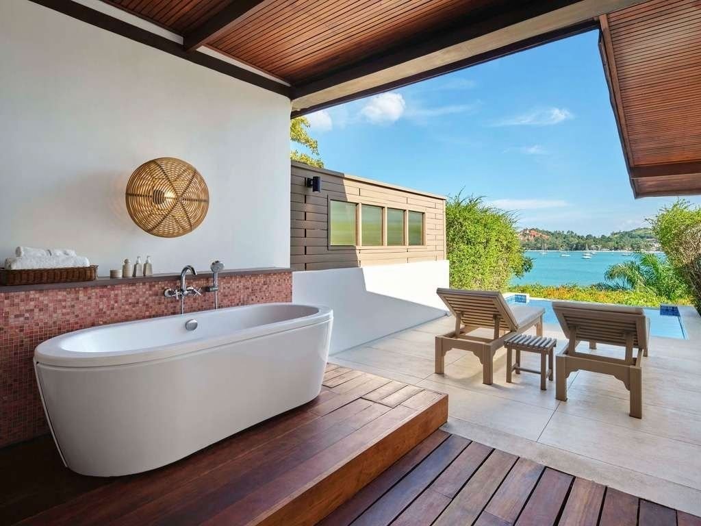 Outdoor bathroom with a white bathtub, two wooden lounge chairs, small table, overlooking a lake with boats, trees, and a partly cloudy sky