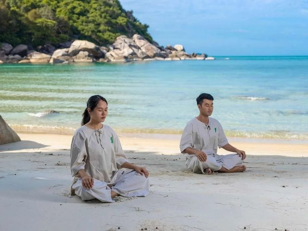 Two people practicing meditation on a sandy beach, sitting cross-legged with eyes closed, near the ocean with a rocky, green-covered island in the background.