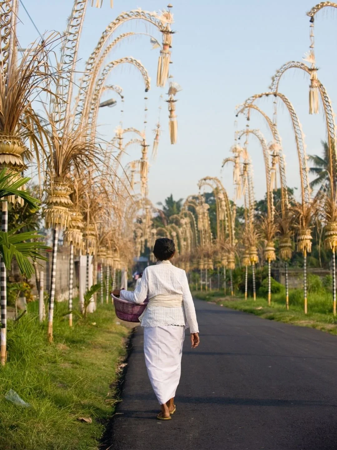 Balinese woman walking along a village road lined with tall ceremonial bamboo penjor decorations in Bali, Indonesia.