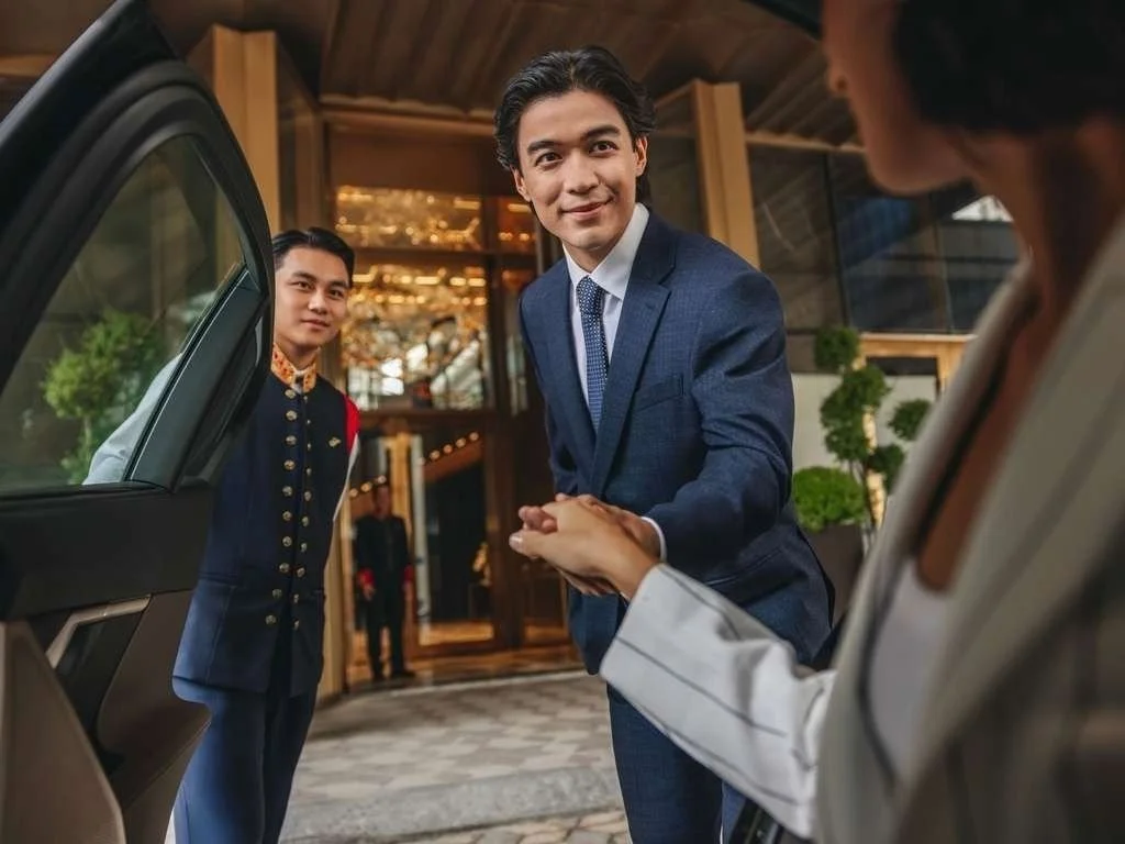 A man in a blue suit is shaking hands with a woman while a bellboy stands nearby outside a hotel entrance.