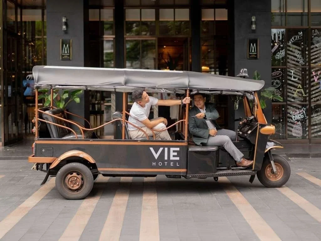 A black tuk-tuk taxi with an open structure parked outside a hotel with a sign that reads 'VIE HOTEL'. The tuk-tuk has a canopy roof and two men sitting inside; one is driving and the other is sitting in the back, reaching out to the driver.