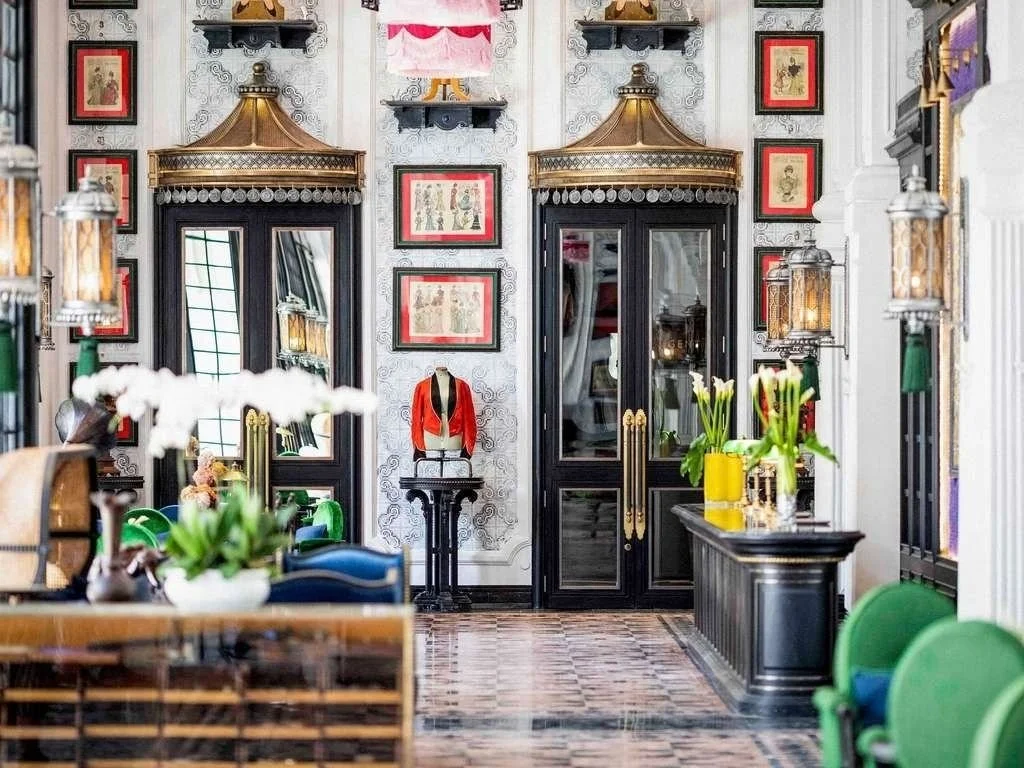 Elegant room with framed artwork, ornate golden canopy, black double doors with gold handles, decorative lanterns, potted orchids, and green upholstered chairs.