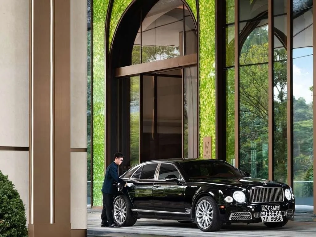 A man in a suit opening the rear door of a black luxury sedan parked in front of a modern building with large glass windows and green vine decorations.