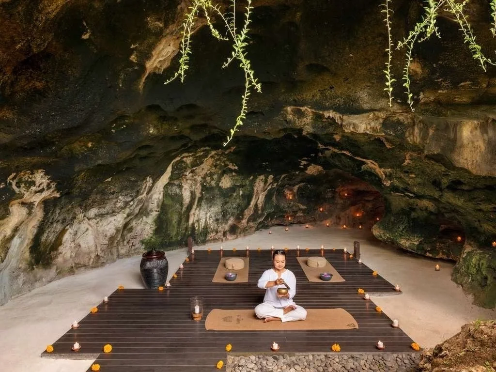 A woman is sitting cross-legged on a mat inside a cave, surrounded by candles and small flower arrangements, with rock formations and hanging plants overhead.