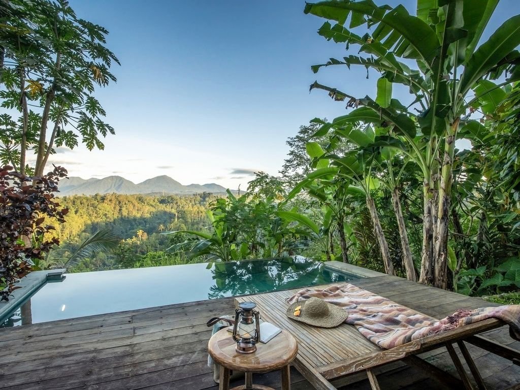 A relaxation area with a wooden deck, striped towel, straw hat, lantern, and soap, overlooking lush greenery and mountains at sunset.