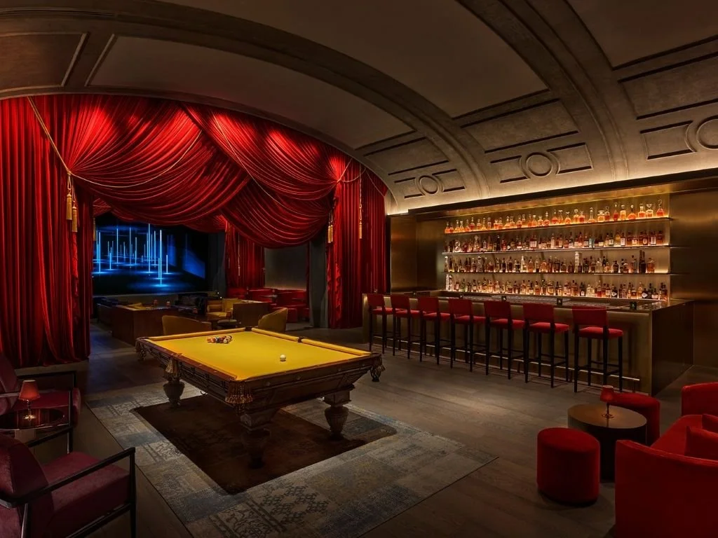 Intimate cocktail lounge featuring dramatic red velvet drapery, amber backlit bar, and custom billiard table at The Singapore EDITION, Singapore.