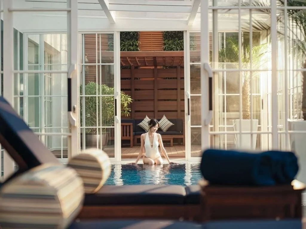 A woman in a white swimsuit kneels in a pool, viewed through glass doors in a sunlit indoor-outdoor pool area with poolside chairs and tropical plants.