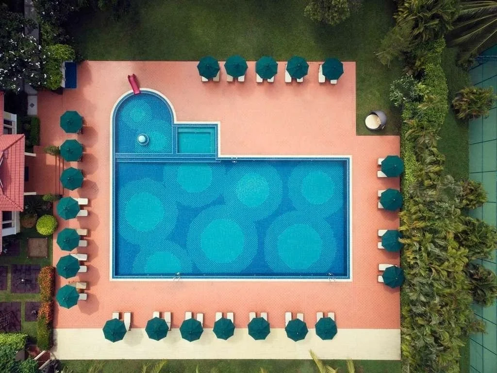 Bird's eye view of a rectangular swimming pool with a small round section at the top left corner, surrounded by green umbrellas and lounge chairs on a brick pool deck, with lush greenery and trees along the edges.