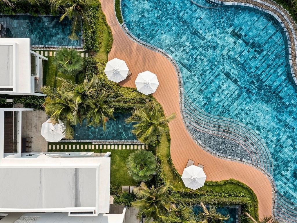 Top-down view of a swimming pool area with curved pool, sun loungers with umbrellas, palm trees, and surrounding greenery and pathways.