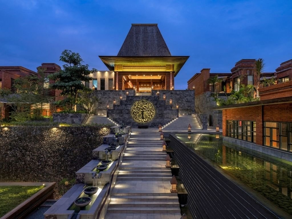 A modern, elegant building with a steep roof, surrounded by stairs, lanterns, and water features, illuminated during dusk.