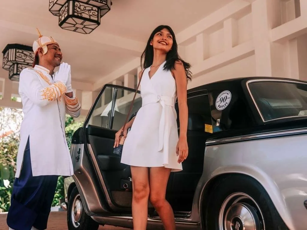 A woman in a white dress standing next to a vintage car, smiling, while a man in traditional attire with a decorative headpiece shows a greeting gesture in front of her.