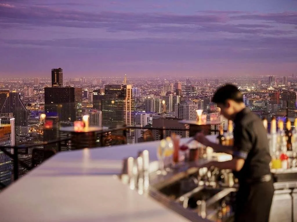 A bartender prepares drinks on a rooftop bar with a city skyline view during dusk, with purple and pink sky and lit-up buildings in the background.