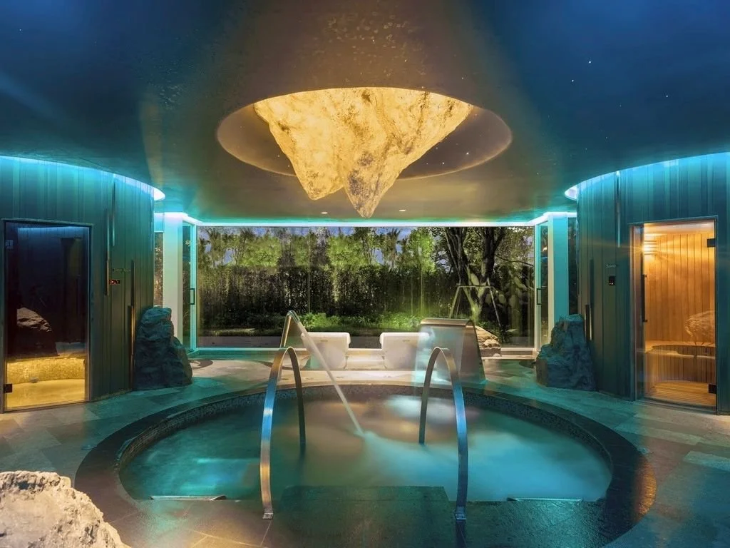 Indoor spa with a whirlpool, two sauna chambers, a large window overlooking trees, and a decorative rock ceiling feature.