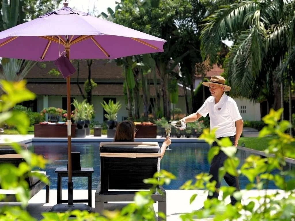 A woman sitting by a pool under a large purple umbrella, being served champagne by a man in a white shirt and wide-brimmed hat.