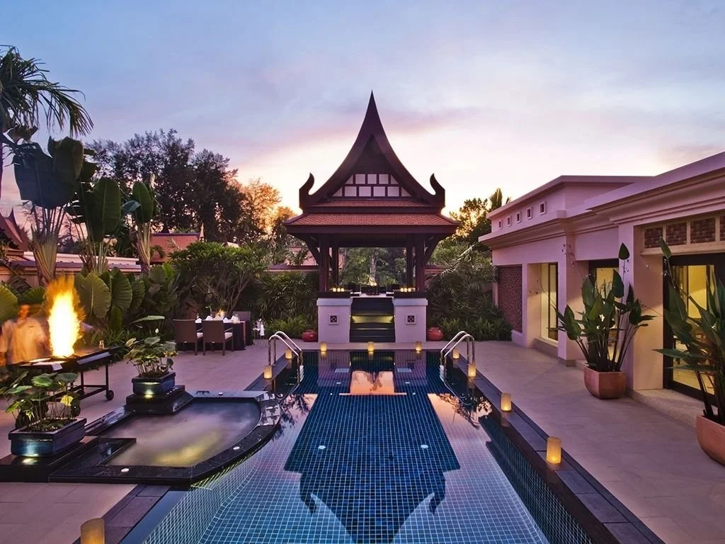 Luxury outdoor pool area with traditional Thai-style pavilion, lush tropical plants, and seating areas at sunset.