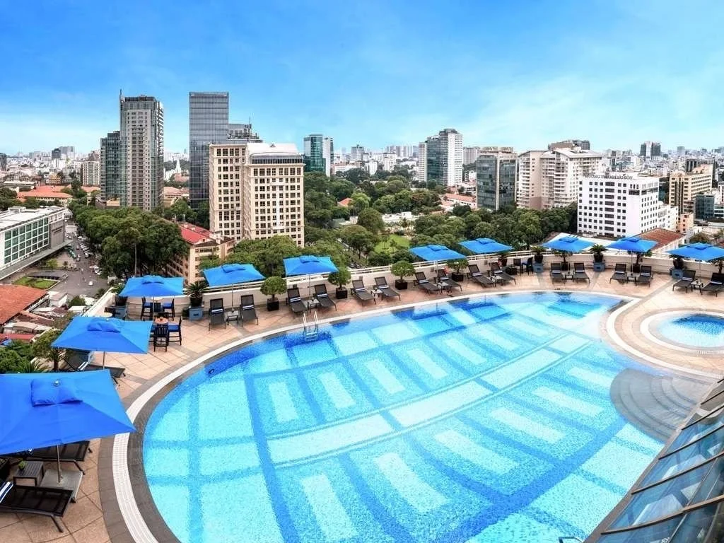 Swimming pool on rooftop with city skyline and tall buildings in the background, blue umbrellas and lounge chairs around the pool.
