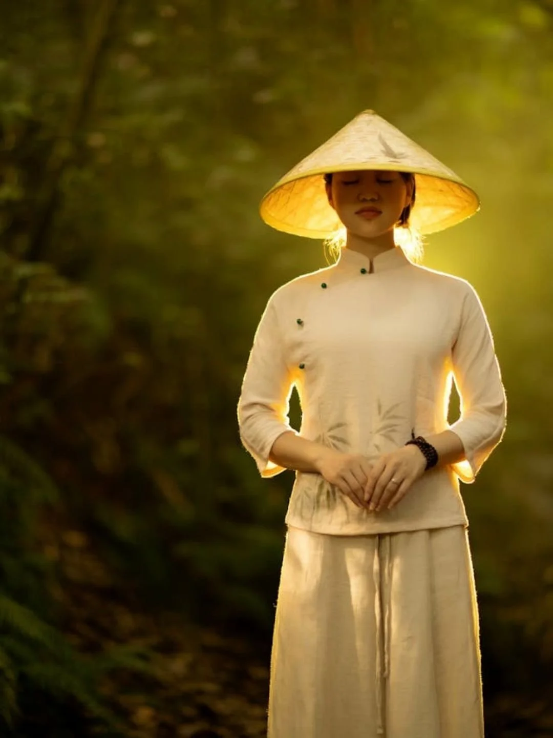 Woman wearing a traditional conical leaf hat (nón lá) bathed in soft morning light amid lush natural surroundings in Vietnam.