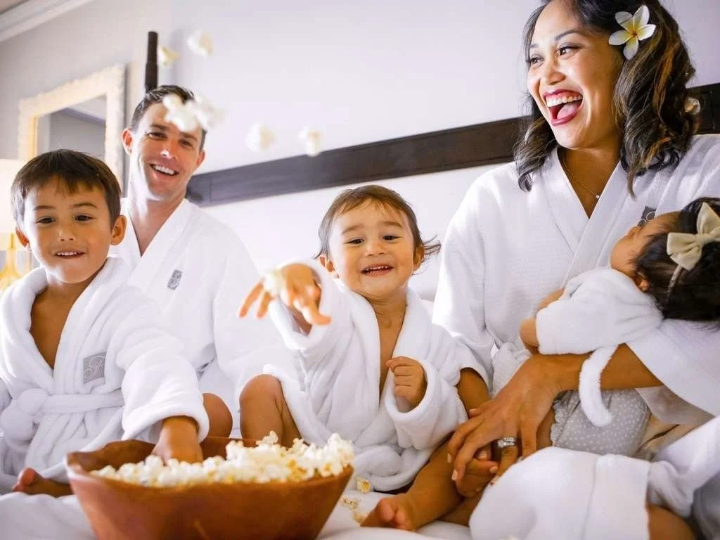 A family of five, dressed in white robes, enjoying a moment together with children laughing and playing, one child reaching for popcorn, in a cozy indoor setting.