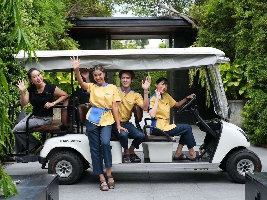 Four smiling people waving from a white golf cart surrounded by greenery.