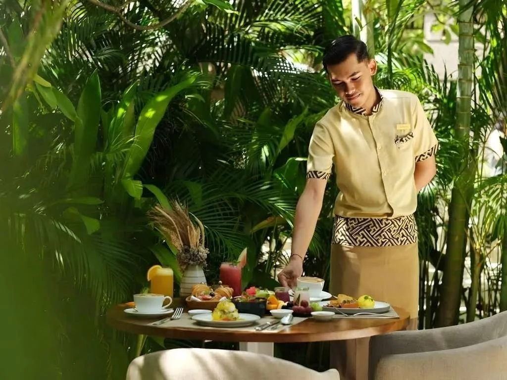 A man in a beige uniform setting a table with breakfast foods and drinks in a lush tropical environment.