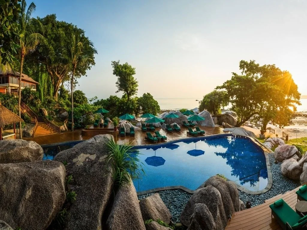 Resort pool area with lounge chairs and umbrellas, surrounded by rocks and trees, overlooking the ocean during sunset.