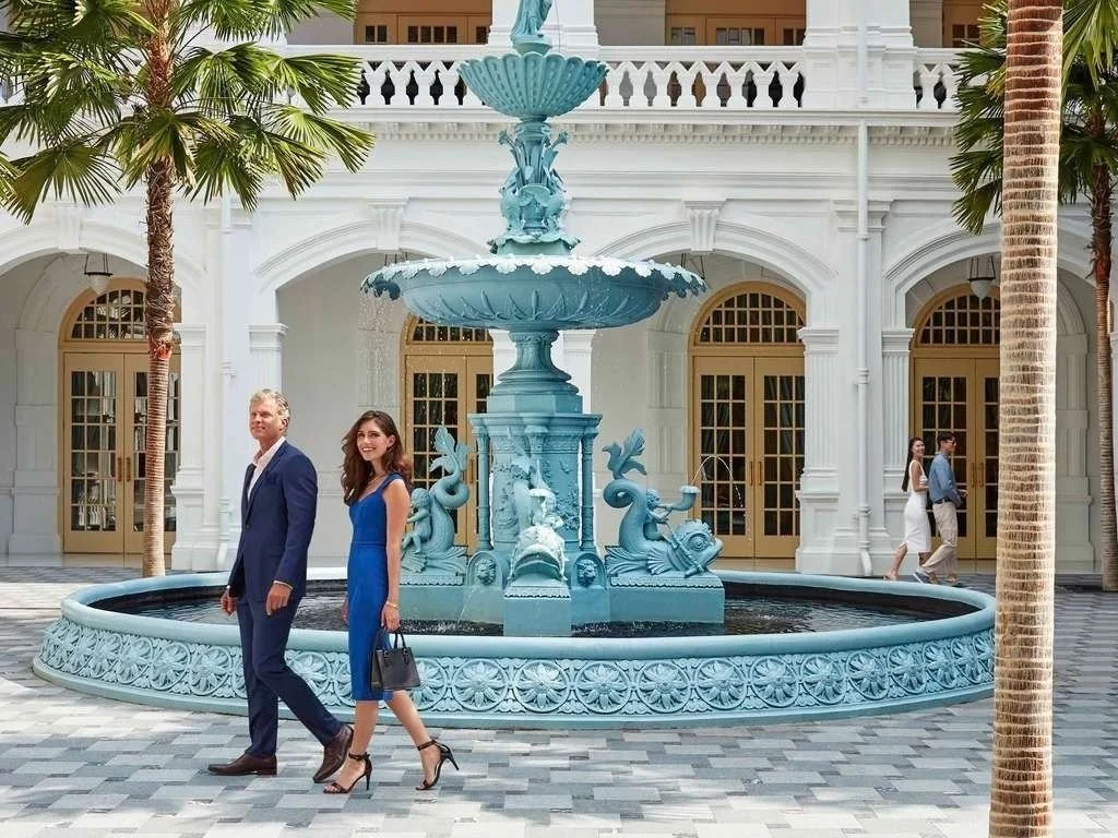 A courtyard with a blue fountain, surrounded by palm trees and a white building with arched doorways. Two people in formal attire walk in the foreground, and a couple in the background walks near the building.
