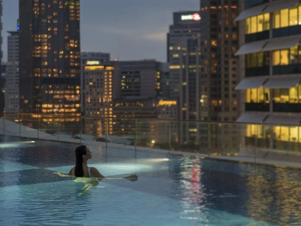 Guest unwinding in the outdoor rooftop pool at twilight against illuminated city skyscrapers at Four Seasons Hotel Kuala Lumpur, Malaysia.