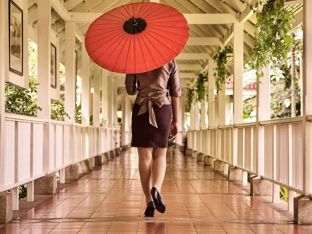 A woman walking through a covered outdoor corridor with white railings, holding a large red umbrella.