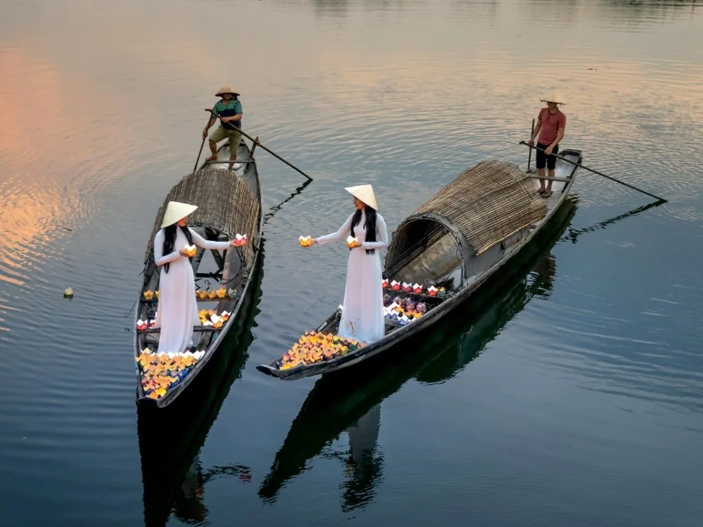 Traditional boats on the river in Hoi An, Vietnam at sunset, with women in white dresses and conical hats holding flower lanterns, creating a serene cultural scene.