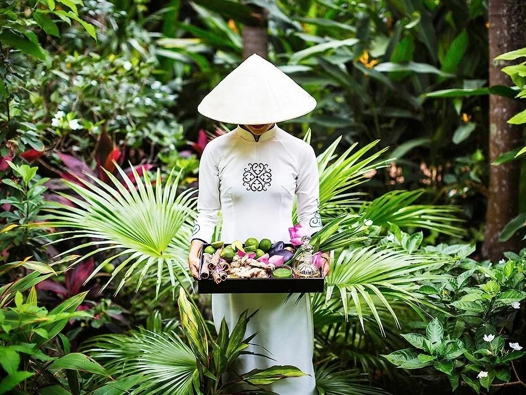 A person dressed in traditional white Vietnamese clothing, wearing a nón lá (conical hat), holding a tray of tropical fruits surrounded by lush green foliage.