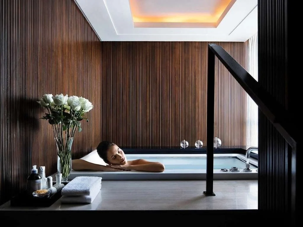 A woman relaxing in a bathtub in a modern bathroom with wood-paneled walls, flower arrangement, towels, and bath accessories.