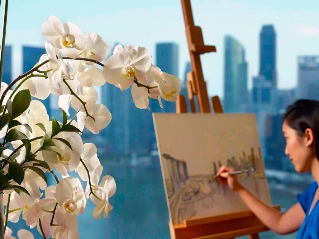 Guest enjoying an in-room art experience at The Ritz-Carlton, Millenia Singapore with orchids and floor-to-ceiling windows overlooking the city skyline.