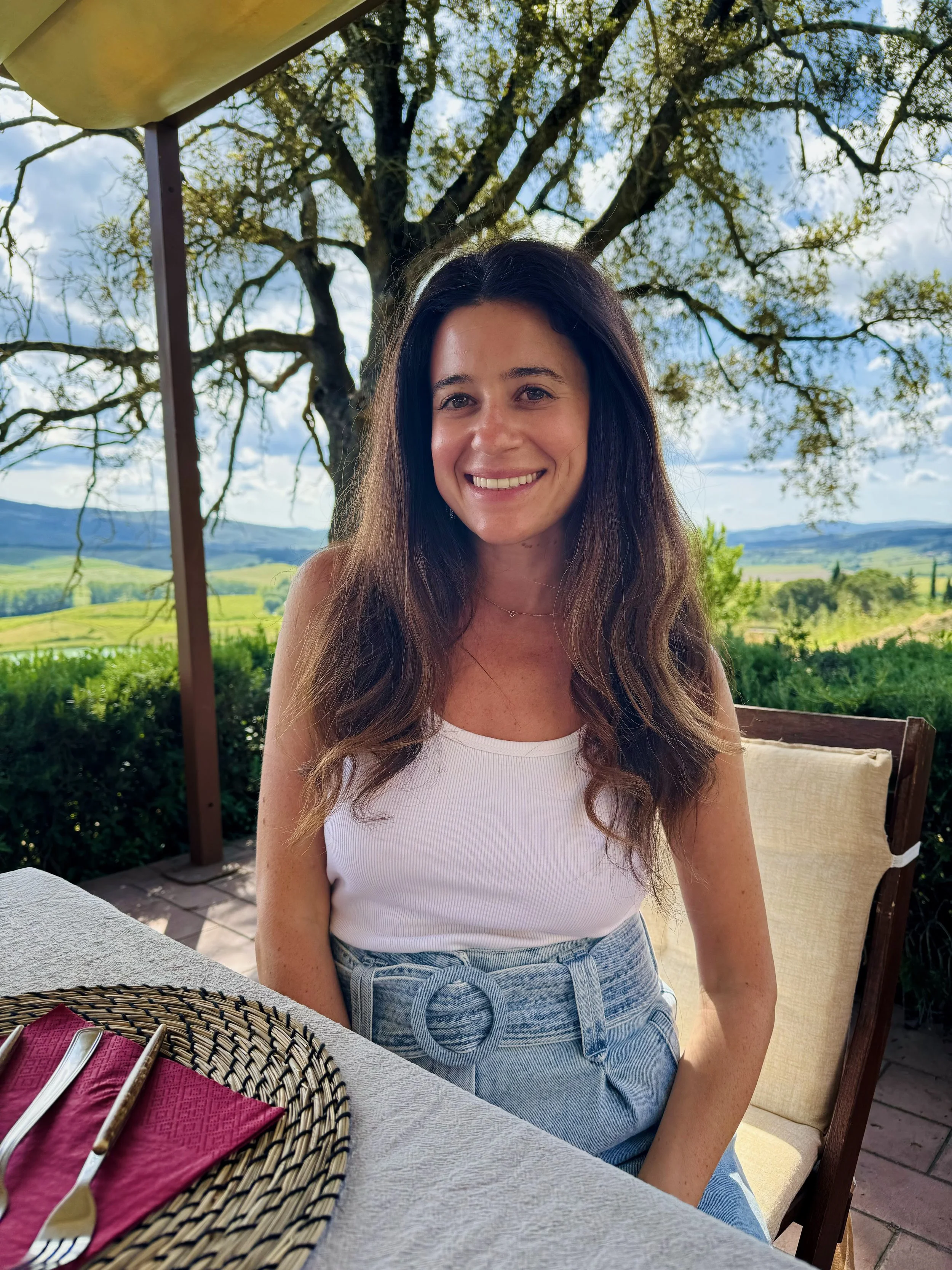 A woman with long brown hair, smiling, wearing a white sleeveless top and high-waisted light blue jeans, sitting outdoors at a dining table with a scenic landscape of green fields, hills, and a tree in the background.