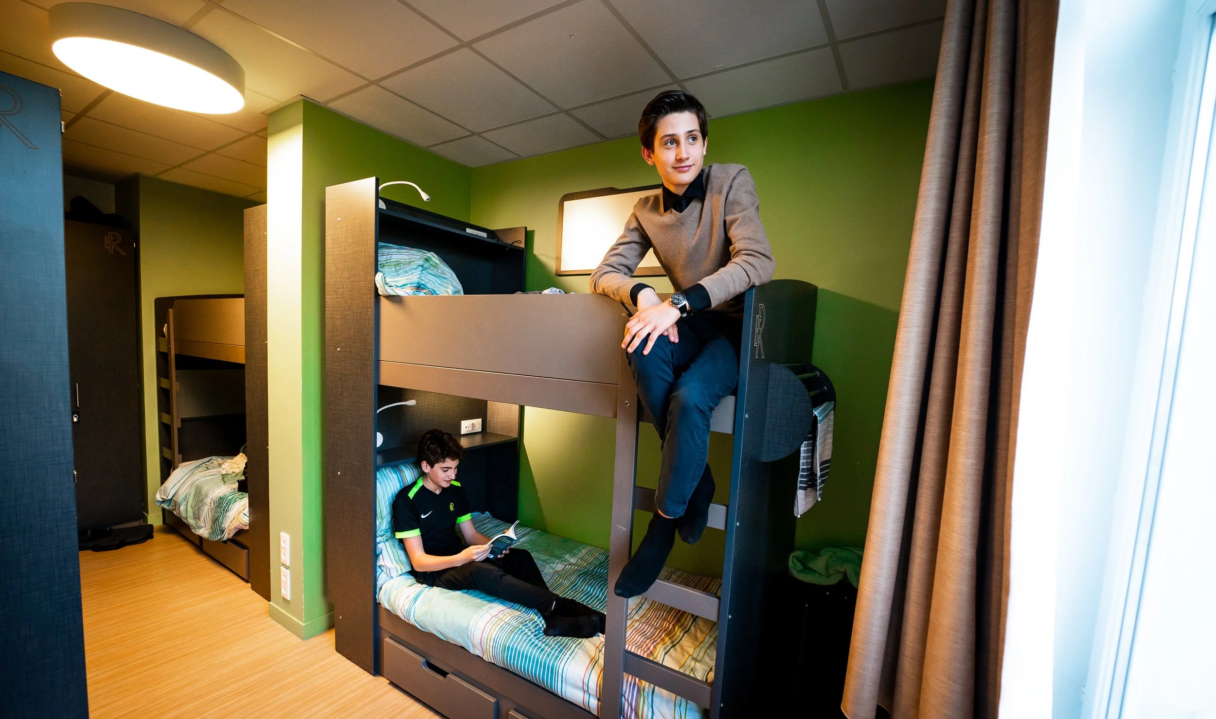 Two boys in a dorm room, one sitting on the bunk bed ladder and the other lying on the bottom bunk reading a book, with green walls and brown curtains.