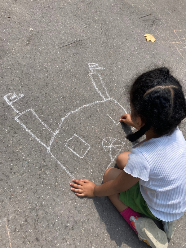 A young girl with braided hair, wearing a white shirt, green shorts, and pink socks, is drawing on the pavement with white chalk. She is creating a drawing of a house with a door, windows, and a chimney, along with a sun in the sky.