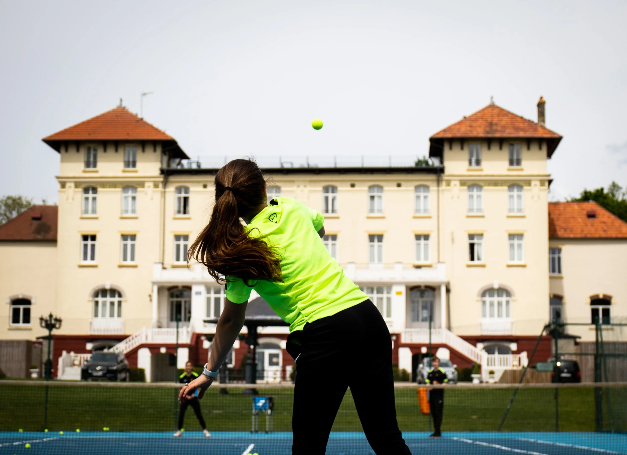 A woman in a neon yellow sports shirt playing tennis on an outdoor court with a large, cream-colored hotel in the background.